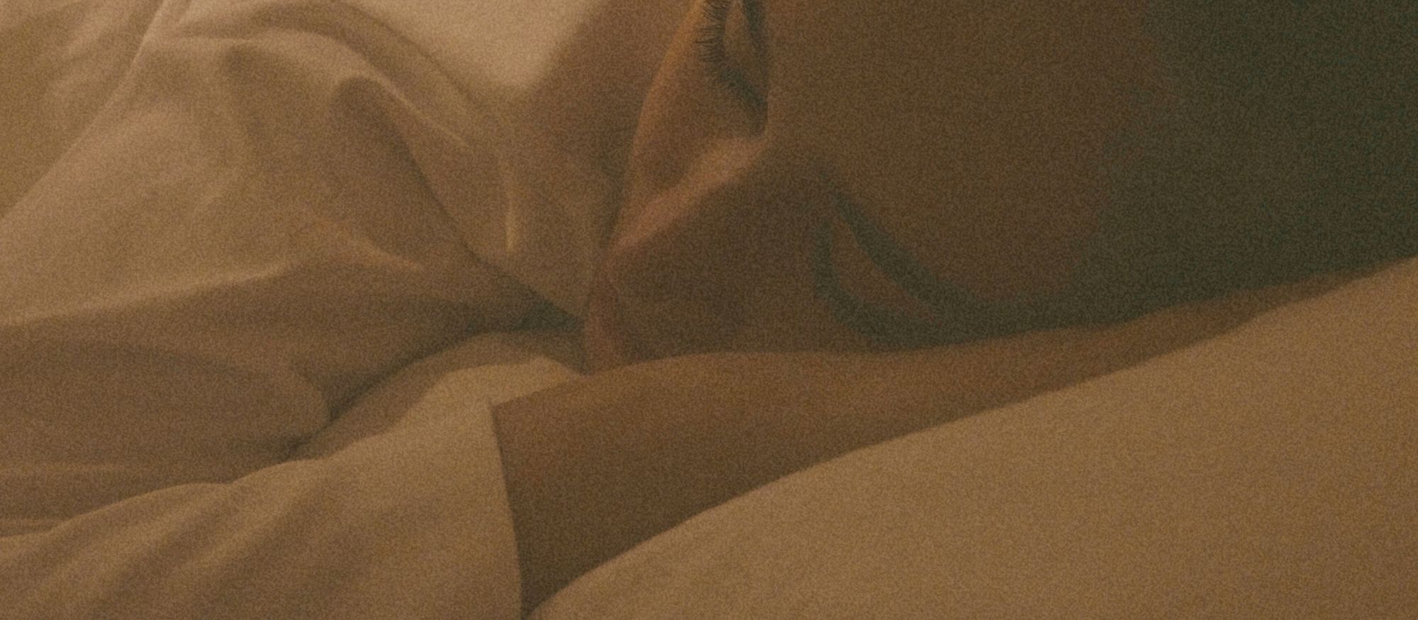 Visage endormi blotti contre l’oreiller, lumière ambrée et draps doux, ambiance calme propice à l’endormissement naturel.