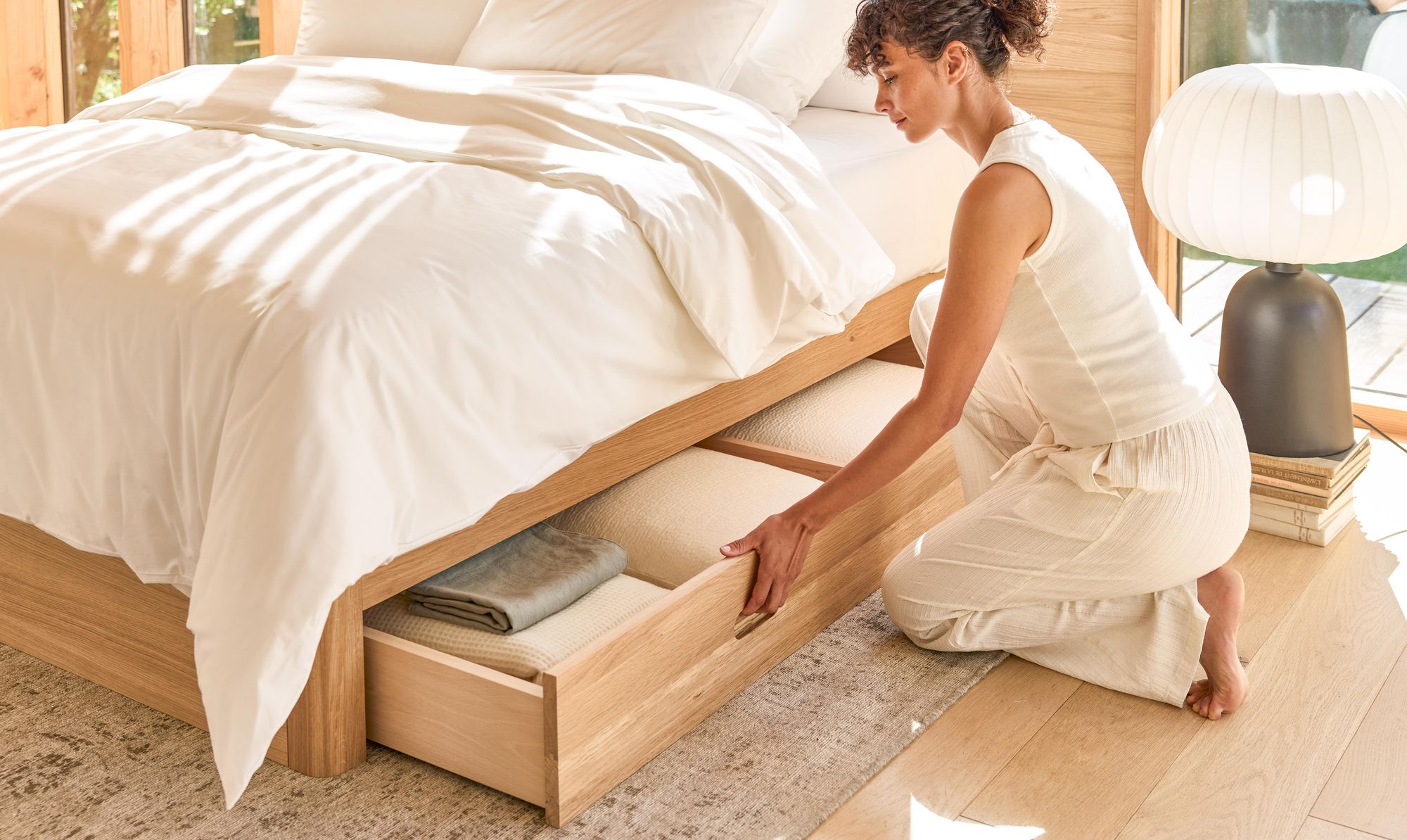 Femme rangeant du linge dans le tiroir d’un lit en chêne massif, chambre lumineuse et ordonnée — solution de rangement intégrée pour une chambre saine.