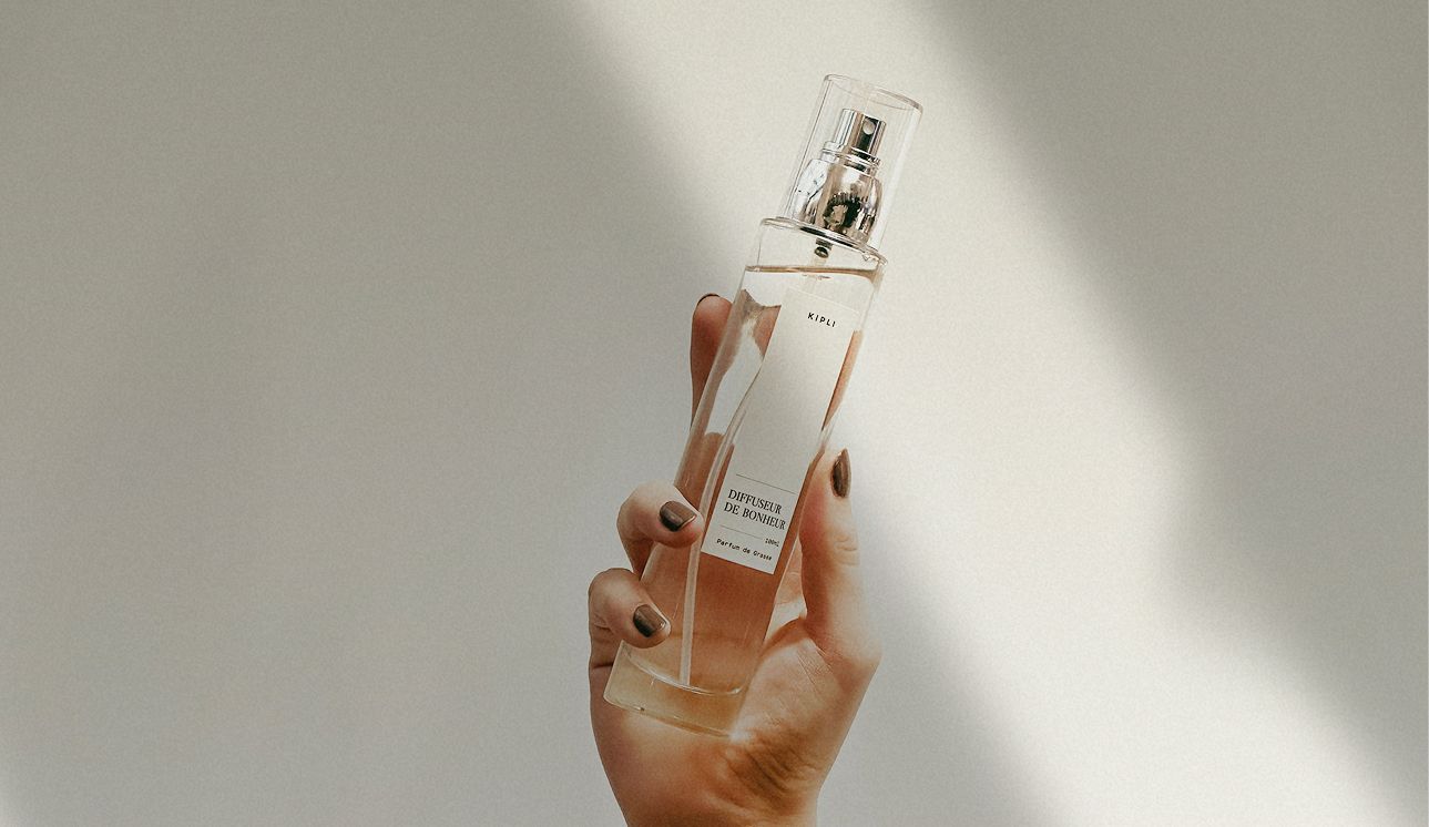 Femme tenant un parfum de maison Kipli dans un flacon en verre ambré, prête à parfumer délicatement une chambre lumineuse pour une atmosphère naturelle et apaisante.
