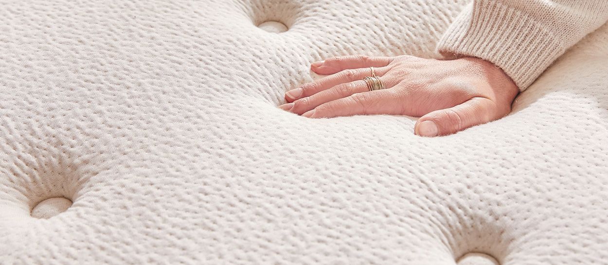 Main appuyée sur la surface matelassée du matelas, léger enfoncement montrant la douceur, la résilience et la respirabilité de l’accueil.