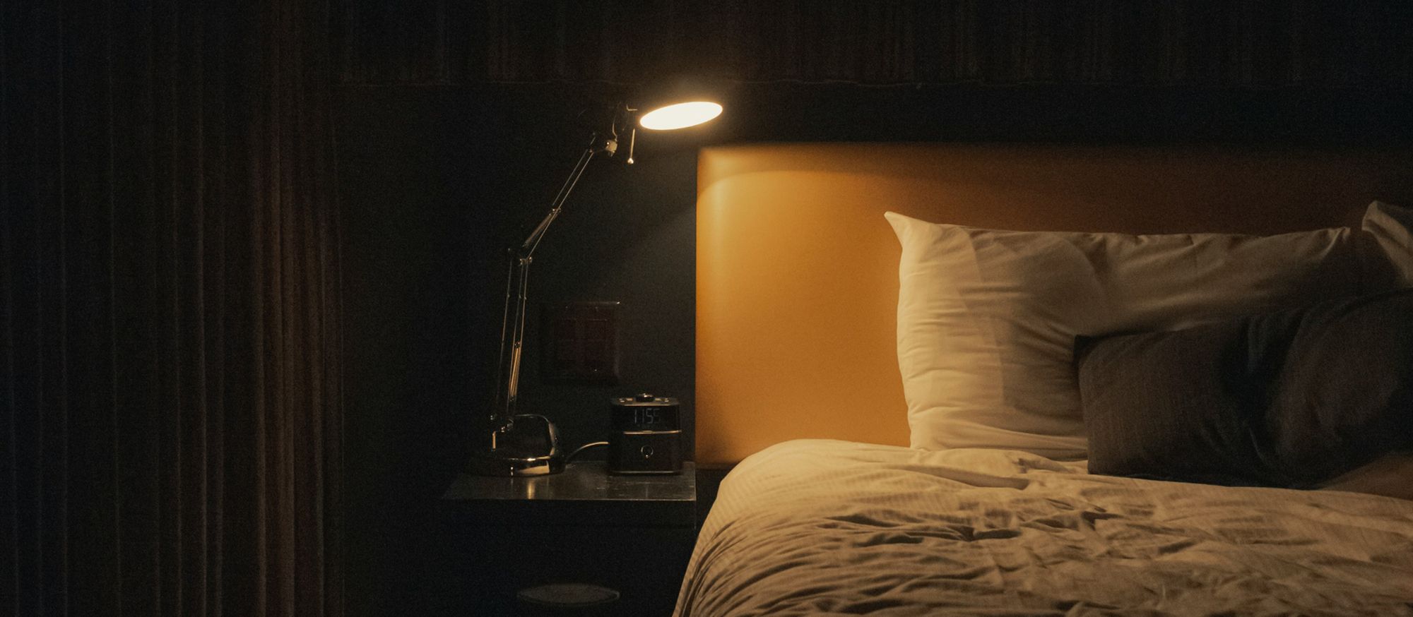 Chambre plongée dans la pénombre, lampe de chevet allumée près d’un réveil numérique, lit prêt pour une nuit paisible.