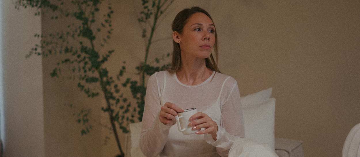 femme assise dans un lit Kipli, enveloppée dans une couette claire, tenant une tasse dans une chambre douce et minimaliste aux tons naturels