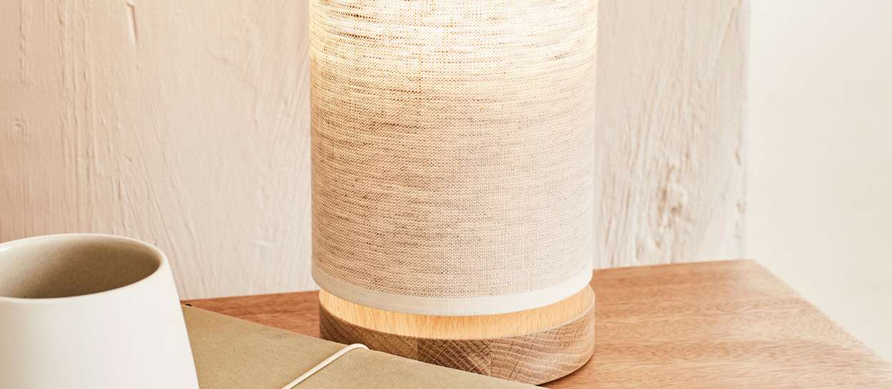 Lampe à poser ISEE en lin naturel avec base en chêne sur une table en bois, diffusant une lumière douce et chaleureuse