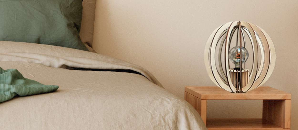 Lampe à poser OYA en lamelles de peuplier sur une table de chevet en bois, à côté d’un lit en linge naturel