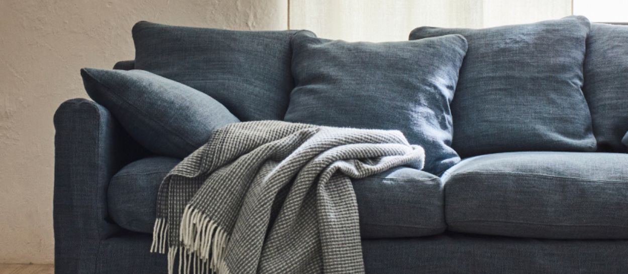 Plaid en laine mérinos GOYA gris à franges posé sur un canapé bleu, parfait pour les soirées d’hiver