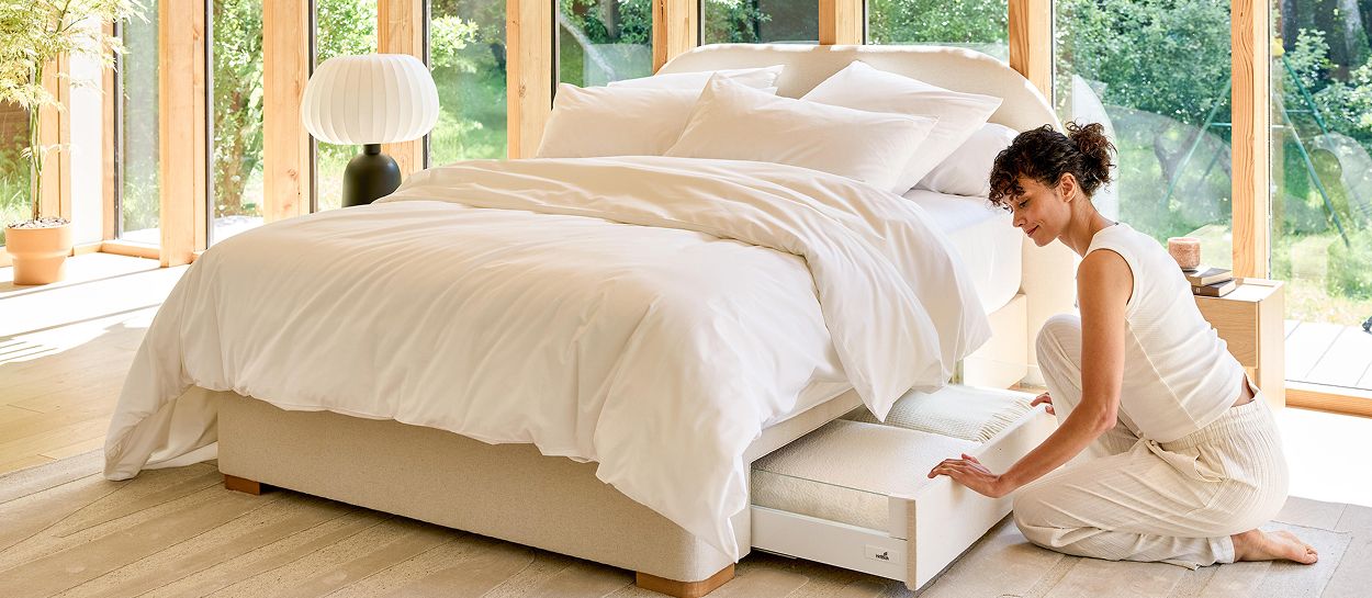 Lit coffre premium La Voile de la collection Au bord du lac : une personne ouvre le rangement intégré au pied du lit, dans une chambre lumineuse et apaisée.