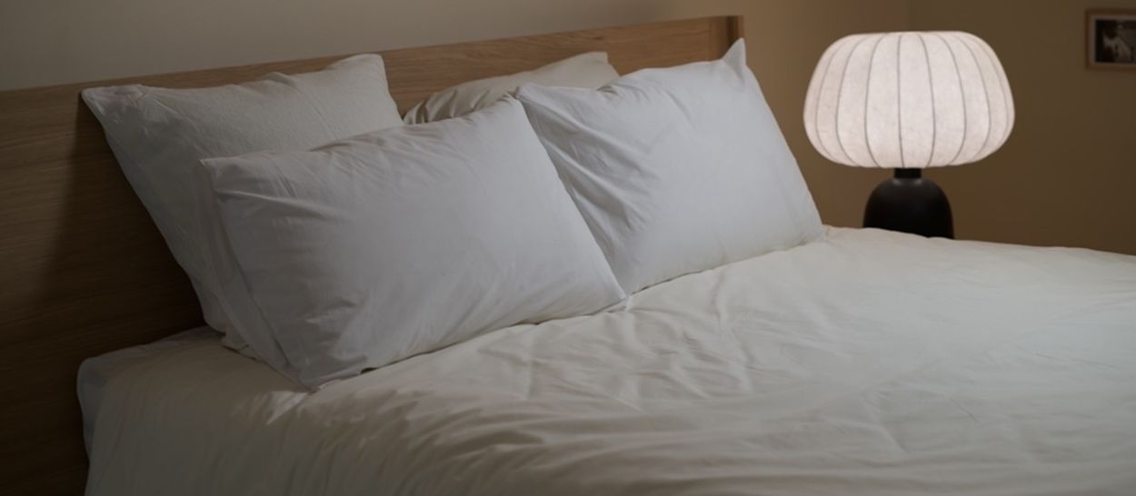 Lit préparé avec draps clairs et coussins en matières naturelles, éclairé par une lampe douce, créant une atmosphère calme et propice au sommeil