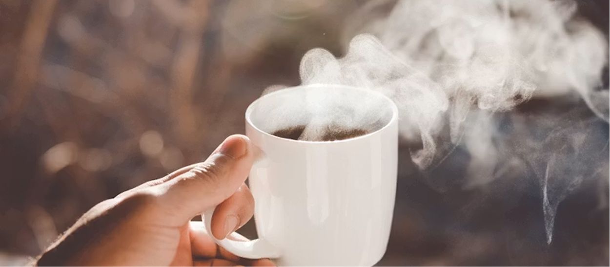 Main tenant une tasse de café chaud fumant, illustrant la surconsommation de caféine chez les télétravailleurs et son impact sur la vigilance nocturne