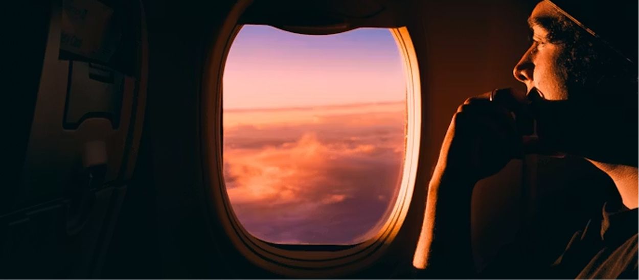 Personne regardant par le hublot d'un avion au coucher de soleil, illustrant l'importance de l'exposition à la lumière pour adapter son horloge biologique avant et pendant un long voyage