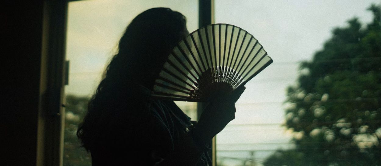 Silhouette d'une femme s'éventant devant une fenêtre en fin de journée, évoquant les bouffées de chaleur et sueurs nocturnes qui perturbent le sommeil à la ménopause