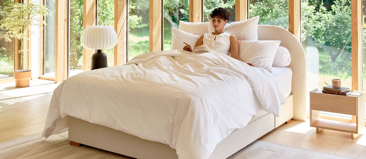 Femme lisant un livre dans un lit Kipli aux draps blancs en coton biologique, près d'une baie vitrée ouverte sur la nature, illustrant un environnement de sommeil sain et de bonnes habitudes pour mieux dormir à la ménopause