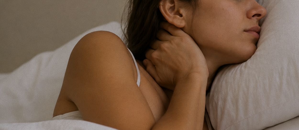 Femme allongée sur le côté portant une main à sa nuque, la tête relevée par un oreiller trop haut, illustrant la tension cervicale et la raideur du cou provoquées par un oreiller mal adapté