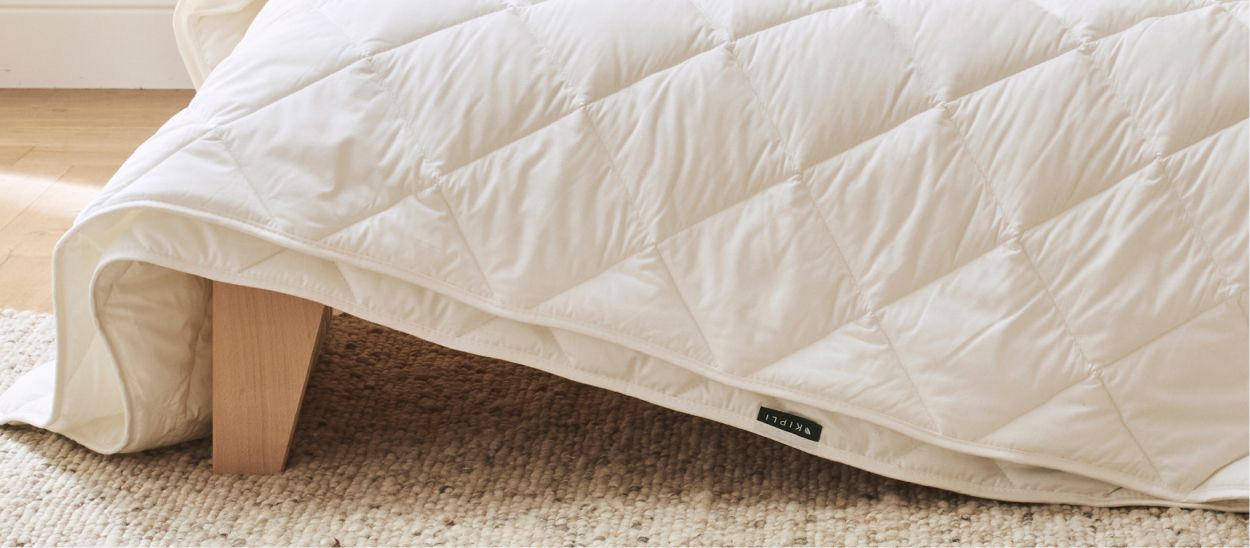 Couette 4 saisons matelassée retombant sur le côté d'un lit en bois clair posé sur un tapis en fibres naturelles, illustrant les différentes configurations d'utilisation de la couette selon la saison et la température de la chambre